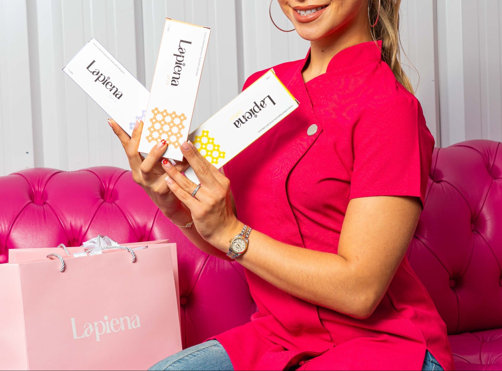 woman in pink holding 3 Lapiena dermal filler product boxes on a pink sofa