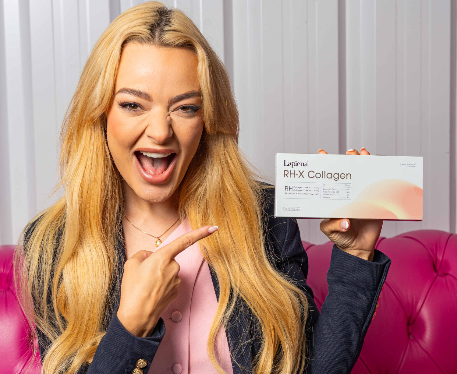 Woman holding a box of of RH-X Collagen Booster and pointing towards it in front of a pink couch.